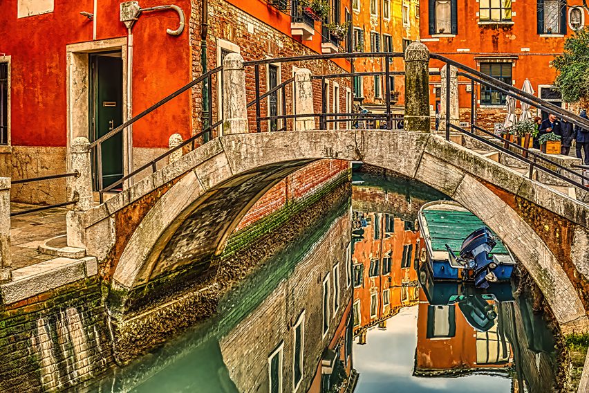 This stunning architectural photograph captures the timeless essence of Venice through a masterfully composed view of a traditional arched stone bridge spanning a quiet Venetian canal, rendered with exceptional artistic precision capturing the luminous interplay of Renaissance architecture, water reflections, and Mediterranean light.